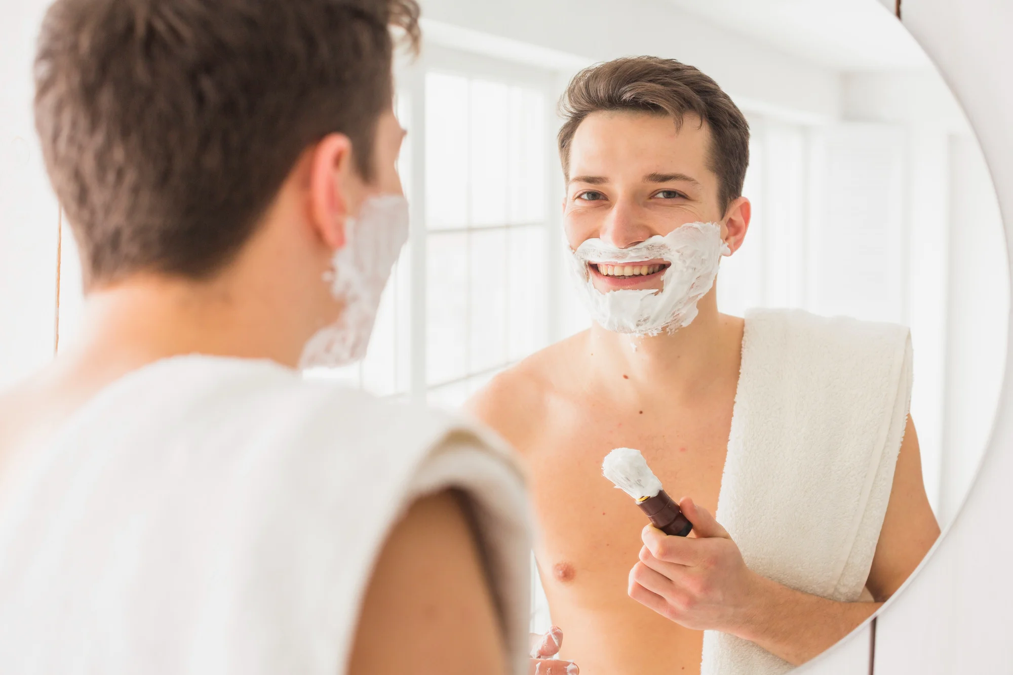 Man shaving with foam using a reusable razor in front of mirror Man shaving with foam using a reusable razor in front of mirror